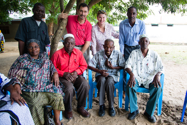 Elders and visitors gather beneath the shade, the portrait carrying both dignity and ease.