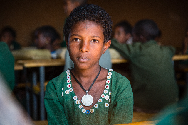 A young student meets the lens with remarkable poise amid the soft blur of her classroom.